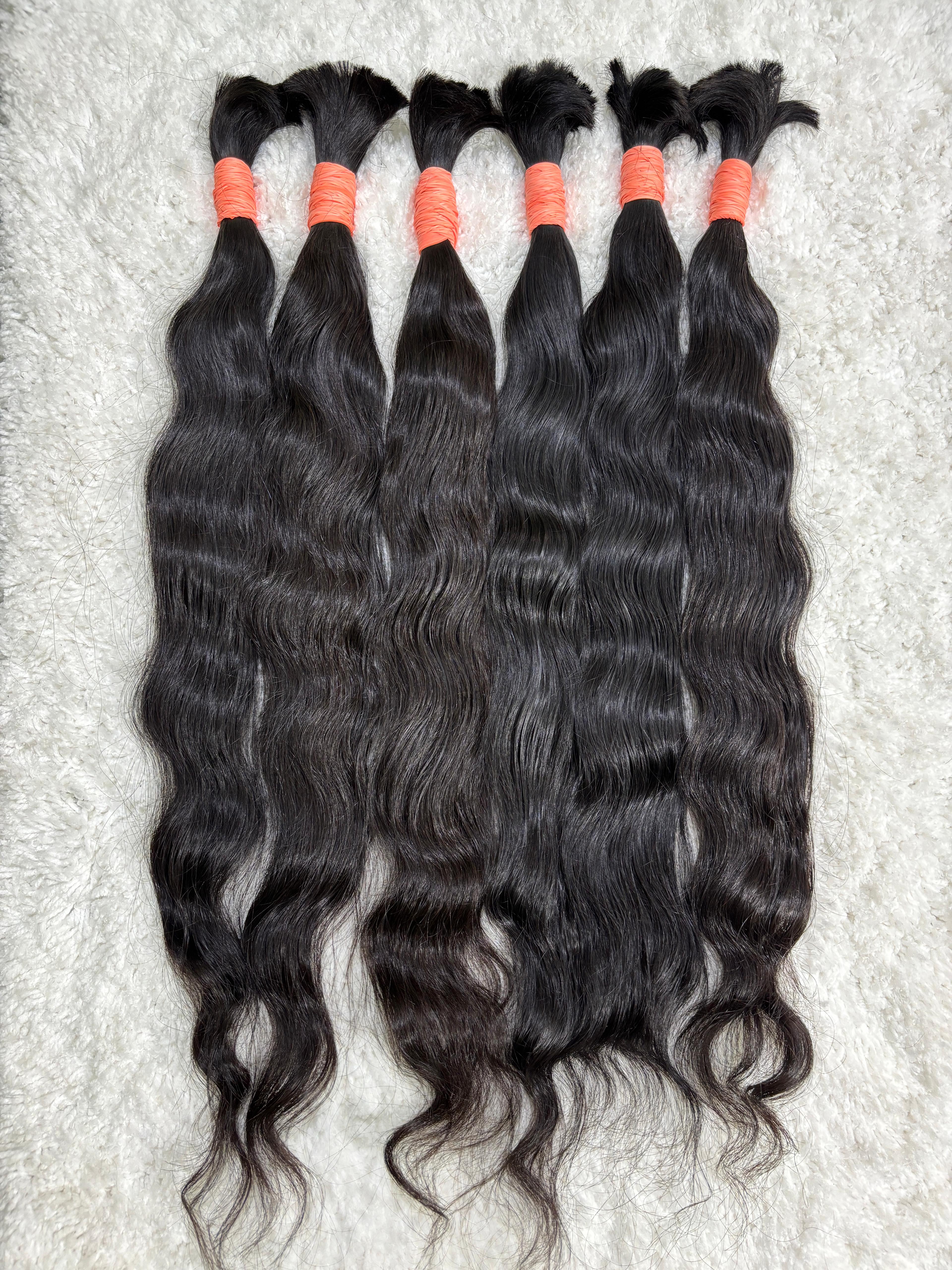 Multiple bundles of raw Indian wavy bulk hair are displayed, tied at the ends with red bands. The hair appears dark and wavy, with a natural texture.